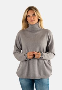 Suéter de cuello alto gris con mangas largas, cuello y puños acanalados, corte relajado y textura suave. Lleva puesto sobre unos pantalones de mezclilla azules.