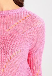 Nahaufnahme eines rosa gestrickten Pullovers mit offenen Strickmustern an Schulter und Ärmel, getragen von einer Person mit braunen Haaren.