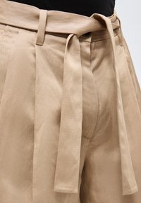 Beige bukser med høj talje med matchende stofbælte bundet i en knude over en sort top, der viser detaljeret talje og folder.