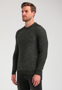 Pull en tricot vert foncé avec un fini texturé, col rond et poignets côtelés. Présente un petit logo sur la zone gauche de la poitrine.