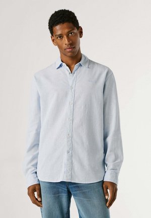 Joven de pie, vistiendo una camisa azul claro con botones y vaqueros azules, frente a un fondo claro y sencillo.