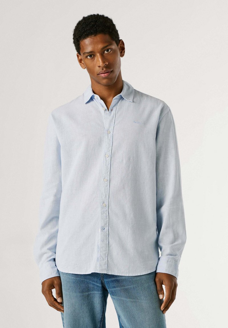 Jeune homme debout, portant une chemise à boutons bleu clair et un jean bleu, sur un fond clair uni.