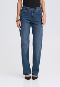 Jean en denim bleu foncé coupe droite, design à cinq poches et fermeture classique à boutons. Porté avec des chaussures noires à bout pointu.