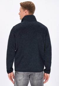 Tumehall fleece-jakk, millel on kõrge kaelus, tekstuuriga kangas ja pikad varrukad. Seljal on sile viimistlus, ilma logode või aktsentideta.