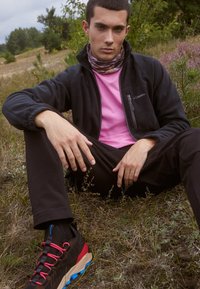 Svart fleecjakke over en rosa skjorte, svarte bukser og fargerike tursko med røde detaljer. Naturlig utendørsmiljø.