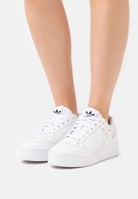 Valkoiset tennarit, joissa on sileä nahkatunnelma, moniväriset niitit sivussa, pyöreä kärki ja paksu kumipohja.