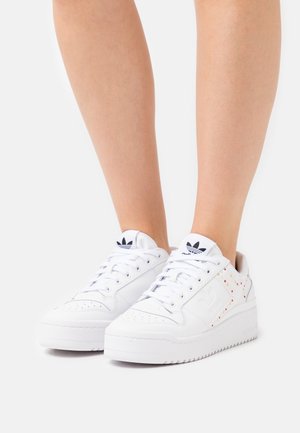 Białe trampki o gładkiej skórzanej teksturze, z wielokolorowymi ćwiekami po bokach, okrągłym noskiem i grubą gumową podeszwą.