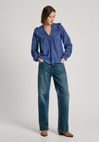 Camicia blu a maniche lunghe con colletto e polsini arricciati, abbinata a jeans scuri a taglio largo e stivali alla caviglia marroni.