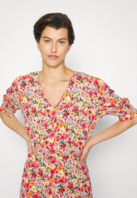 Vestido floral com mangas curtas em balão, decote em V e padrão floral em rosa, amarelo e verde sobre um fundo claro, feito de tecido leve.