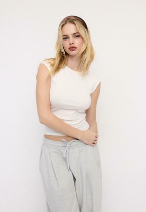 Giovane donna con capelli biondi che indossa un top bianco senza maniche e pantaloni della tuta grigi, in piedi davanti a uno sfondo bianco uniforme.