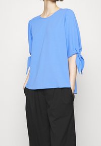 Top azzurro a maniche corte con scollatura rotonda, tessuto a trama e dettagli con laccio sulle maniche, abbinato a pantaloni neri dal taglio ampio.