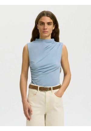 Femme aux longs cheveux bruns porte un haut bleu clair sans manches et un pantalon beige avec une ceinture marron, debout avec une main dans la poche.
