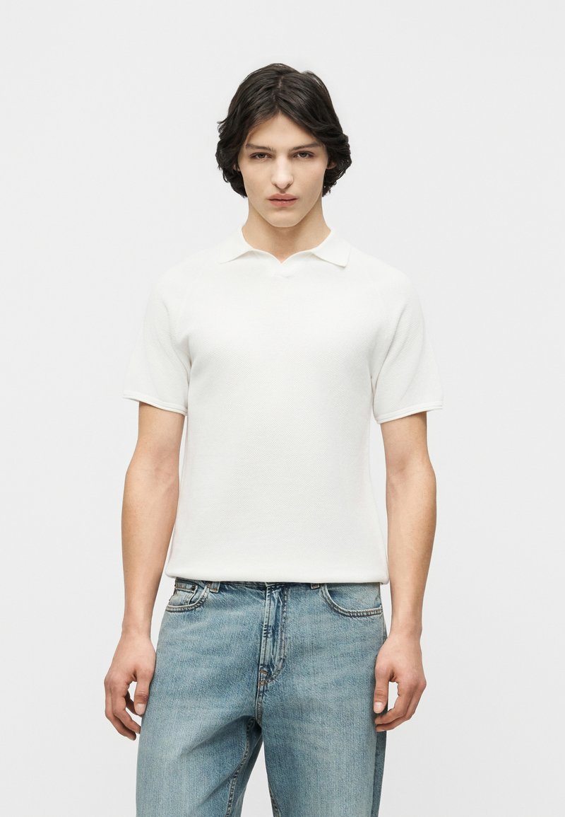 Jeune homme aux cheveux bruns mi-longs portant un polo blanc à manches courtes et un jean bleu clair, debout devant un fond uni.