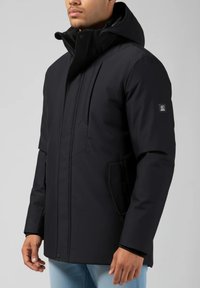 Man som bär en svart vinterjacka med huva, blixtlås och knappar, står mot en enfärgad grå bakgrund.