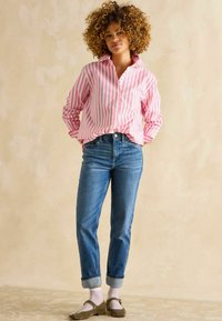 Rosa und weiße gestreifte Hemd mit Knöpfen, blaue Jeans und olivgrüne Schuhe mit weißen Punkten. Hochgekrempelte Ärmel und Manschetten.