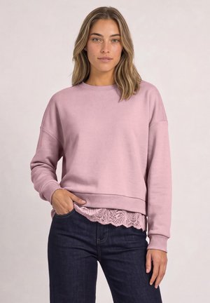 Une femme porte un sweat-shirt rose par-dessus un haut bordé de dentelle et un jean foncé, une main dans la poche.