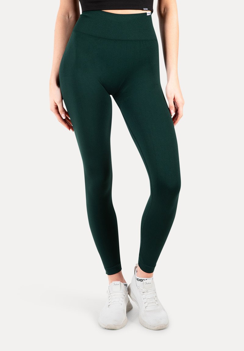 Ciemnozielone legginsy z wysokim, prążkowanym paskiem w talii, wykonane z elastycznego materiału, w połączeniu z białymi butami sportowymi.