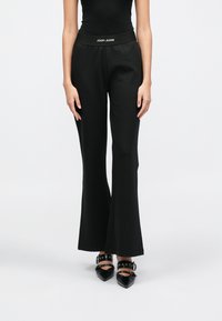 Calças pretas com corte evasê, textura suave e cintura elástica com a inscrição "JOOP! JEANS" em branco, combinadas com sapatos pretos pontiagudos.