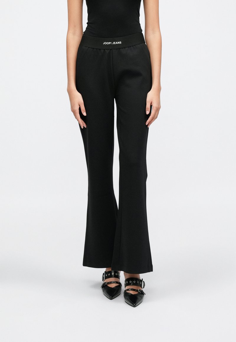 Calças pretas com corte evasê, textura suave e cintura elástica com a inscrição "JOOP! JEANS" em branco, combinadas com sapatos pretos pontiagudos.