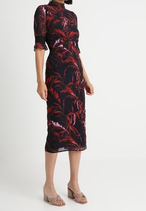 Robe midi fleurie noire avec des motifs de feuilles rouges et roses vibrants, manches courtes transparentes et silhouette ajustée. Associée à des talons blocs marron clair.