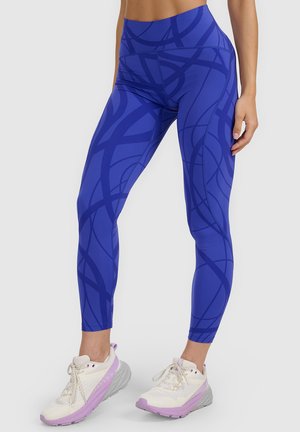 Niebieskie legginsy z abstrakcyjnymi ciemnoniebieskimi liniami noszone przez stojącą osobę, w połączeniu z białymi i jasnofioletowymi butami sportowymi.