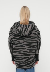 Veste en moumoute à motif zèbre en gris et noir, avec une capuche, coupe oversized et poignets en côte noir. Tissu en polaire doux et texturé.