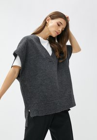 Grijze gebreide oversized vest met een V-hals, geribbelde randen en ruwe zomen, gedragen over een wit shirt. Donkere broek maakt de outfit compleet.