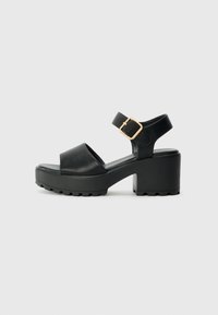 Schwarze Plattformsandalen mit einer glatten Lederoberfläche, breiter Vorderstrap, verstellbarem Knöchelriemen mit einer goldenen Schnalle und strukturiertem Sohlen.