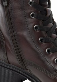 Bottine en cuir marron avec lacets noirs, œillets métalliques, une fermeture éclair sur le côté et une semelle noire texturée. Fini lisse avec des coutures subtiles.