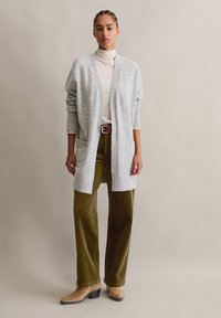 Grauer, langer Cardigan mit offenem Vorderteil und Taschen, getragen �über einem karierten Rollkragenpullover, kombiniert mit olivgrünen Cord-Hosen und beigen Stiefeln mit Absatz.