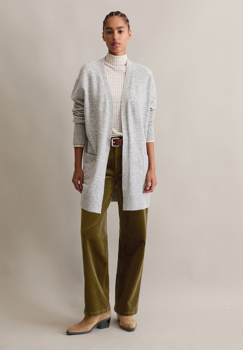Grauer, langer Cardigan mit offenem Vorderteil und Taschen, getragen über einem karierten Rollkragenpullover, kombiniert mit olivgrünen Cord-Hosen und beigen Stiefeln mit Absatz.