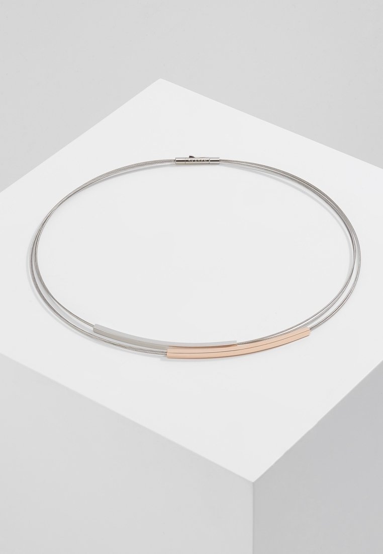 Collier ras-du-cou minimaliste avec des fils d'argent fins et deux barres métalliques incurvées, une argentée et une en or rose, exposé sur un socle blanc.