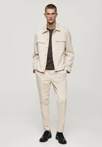 Beige jacket with large chest pockets and beige pants, paired with a dark t-shirt and black shoes. Soft fabric, tailored fit.