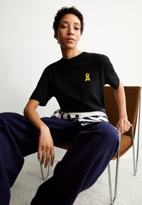 Zwarte t-shirt met geel lintlogo, ontspannen pasvorm en ronde hals, gecombineerd met losse marineblauwe broek. Model zit op een stoel.