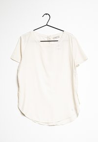 T-Shirt-Bluse aus leichtem Creme-Stoff, mit rundem Ausschnitt, geschwungenem Saum, seitlichen Schlitzen und einem kleinen Metallsignet in der Nähe des unteren Saums.