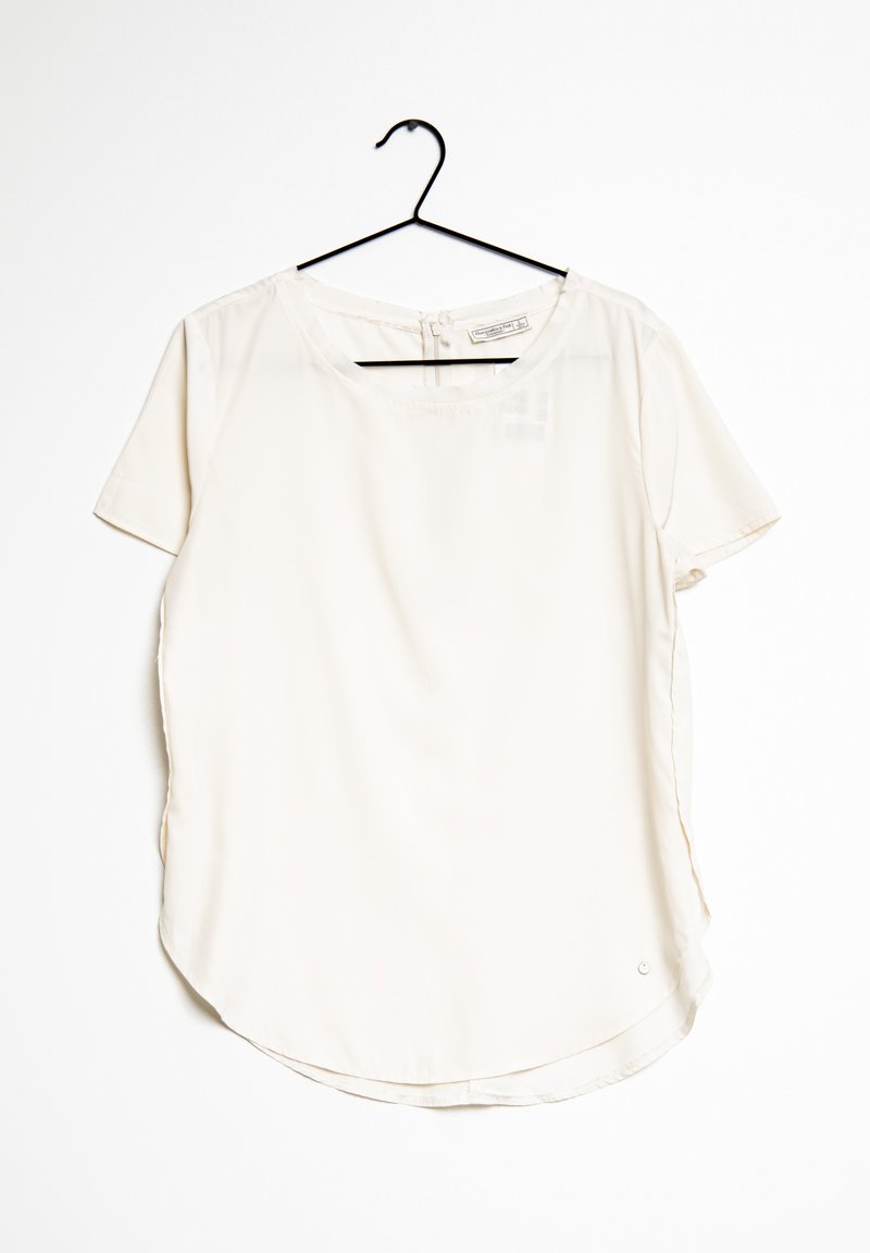 T-Shirt-Bluse aus leichtem Creme-Stoff, mit rundem Ausschnitt, geschwungenem Saum, seitlichen Schlitzen und einem kleinen Metallsignet in der Nähe des unteren Saums.