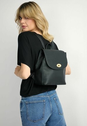 Kobieta o blond włosach, ubrana w czarną bluzkę i niebieskie dżinsy, niosąca na plecach czarny skórzany plecak o fakturze z złotym zapięciem.