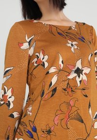 Hnědý dlouhý rukáv svetr s květinovým vzorem v růžové, bílé a modré. Má kulatý výstřih a hladkou, jemnou strukturu.