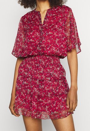 Femme portant une robe courte rouge à fleurs avec des manches évasées transparentes et une taille smockée, debout les bras détendus le long du corps.