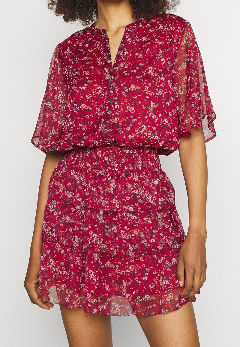 Femme portant une robe courte rouge à fleurs avec des manches évasées transparentes et une taille smockée, debout les bras détendus le long du corps.