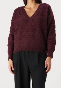 Vinröd stickad tröja med V-ringning, med texturerade mönster och ribbade muddar. Kombinerad med svarta byxor, som framhäver en mysig stil.