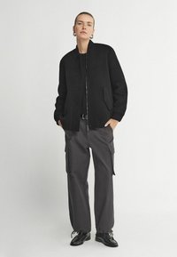 Svart bomberjacka med ribbad krage och två sidofickor, i kombination med grå cargobyxor med sidofickor, allt bärs med svarta skor.