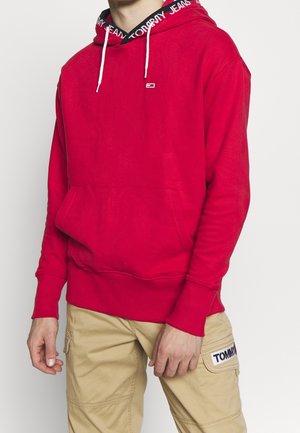 Osoba w czerwonej bluzie z kapturem z napisem "Tommy Jeans" na podszyciu kaptura oraz beżowych spodniach typu cargo z naszywką Tommy na kieszeni.