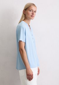 Lichtblauwe t-shirt met korte mouwen en een ronde halslijn, vijfknopenlijst en een zachte, gladde textuur. Gecombineerd met witte broek.
