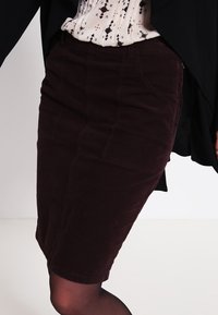 Burgunderfarbene Kordrock im Bleistiftschnitt mit mittellanger Länge, ausgestattet mit seitlichen Taschen und sichtbaren Nähten. Glatter Stoff, figurbetontes Design.