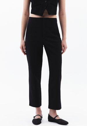 Femme portant un pantalon noir court, un gilet noir sans manches boutonné et des chaussures plates noires, debout sur fond blanc.