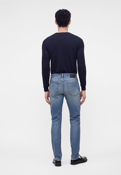 Homme debout de dos, portant une chemise noire à manches longues, un jean bleu et des chaussures noires sur un fond blanc uni.