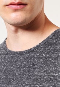 Close-up van het ondergedeelte van het gezicht van een persoon, de nek en de bovenste borst die een grijze textuurtrui met ronde hals draagt.