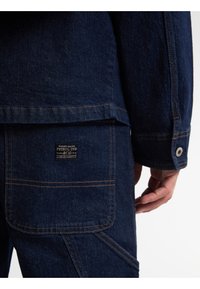 Mørkeblå denimjakke med orange syninger, en lomme med et mærkelogo og en knaplukning på ærmet.