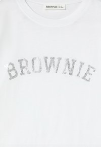 Camiseta de algodón blanca con la palabra "BROWNIE" en texto de purpurina plateada, con fuente redondeada, cuello redondo y mangas cortas.